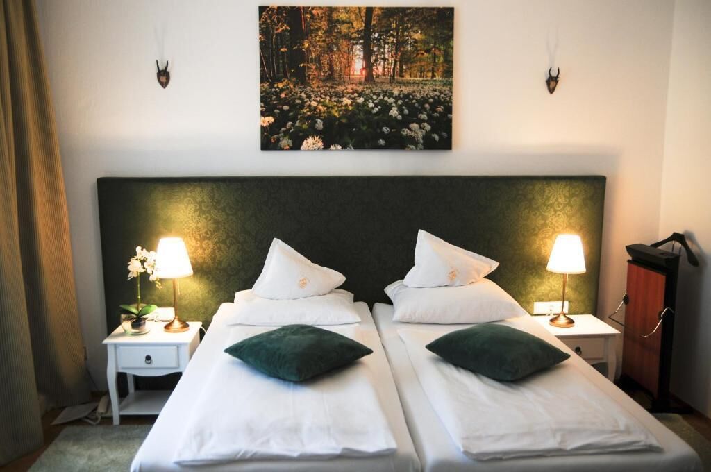 Habitación doble con dos camas, cada una con una almohada y una lámpara. En la pared, sobre las camas, hay un cuadro de un bosque.