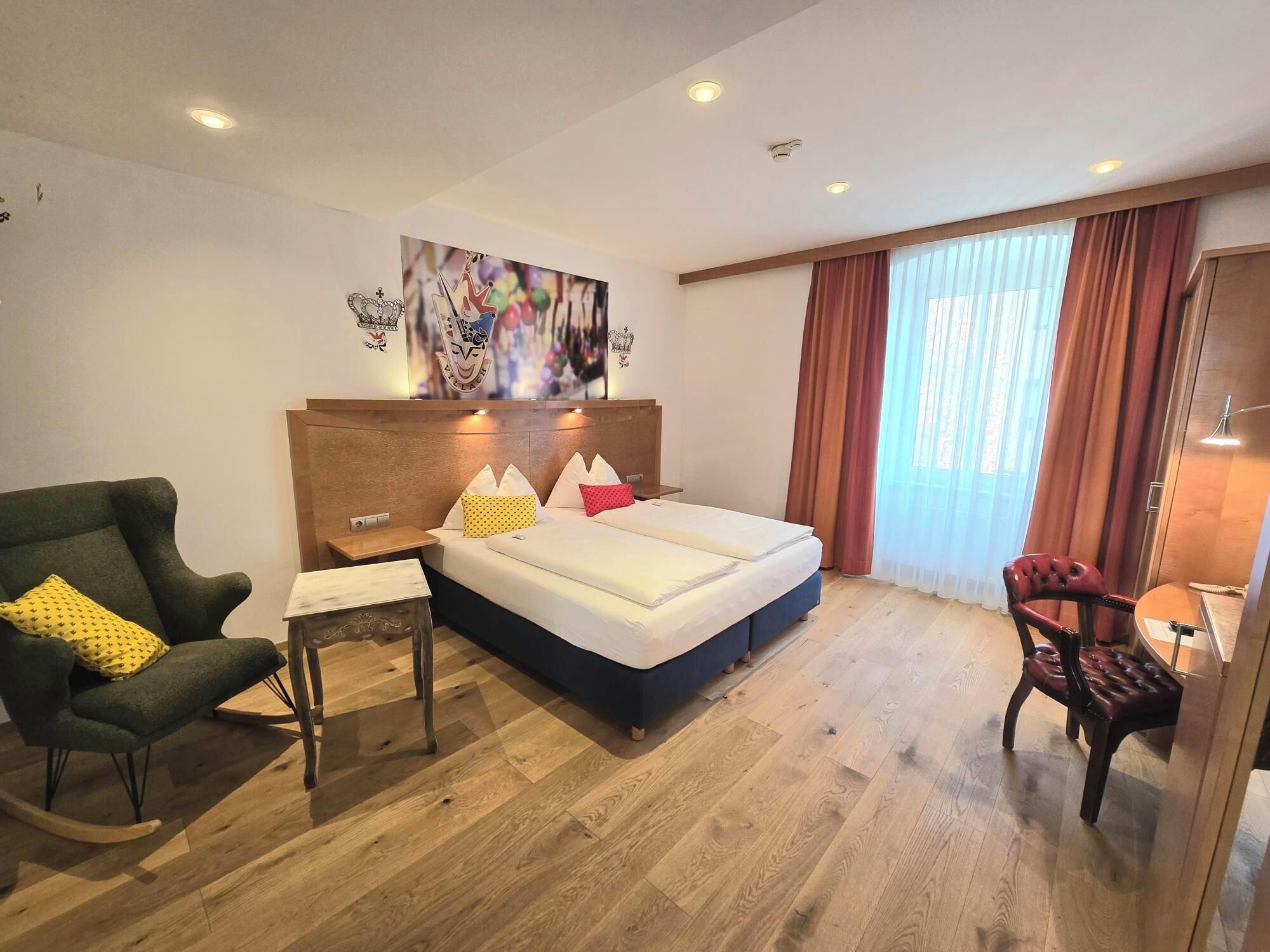 Una camera d'albergo con un letto, una scrivania, una sedia e un'ampia finestra con tende.