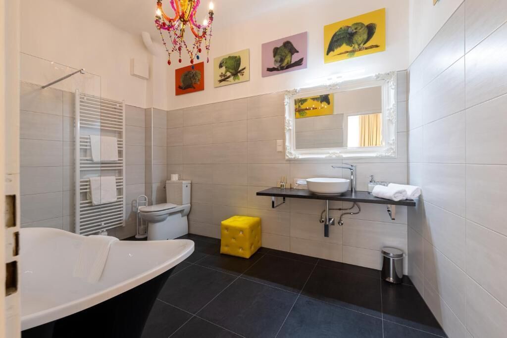 A modern bathroom with a bathtub, toilet, and sink. There are wall-mounted shelves and a decorative chandelier.