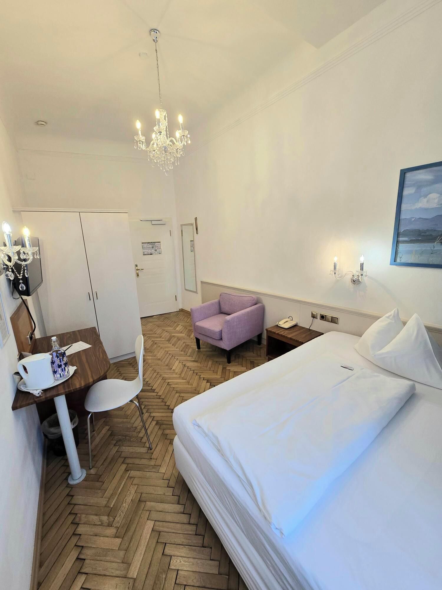 Habitación de hotel con cama, escritorio y silla, sillón púrpura, candelabro y un cuadro en la pared.