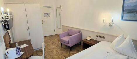 Habitación de hotel con cama, escritorio y silla, sillón púrpura, candelabro y un cuadro en la pared.