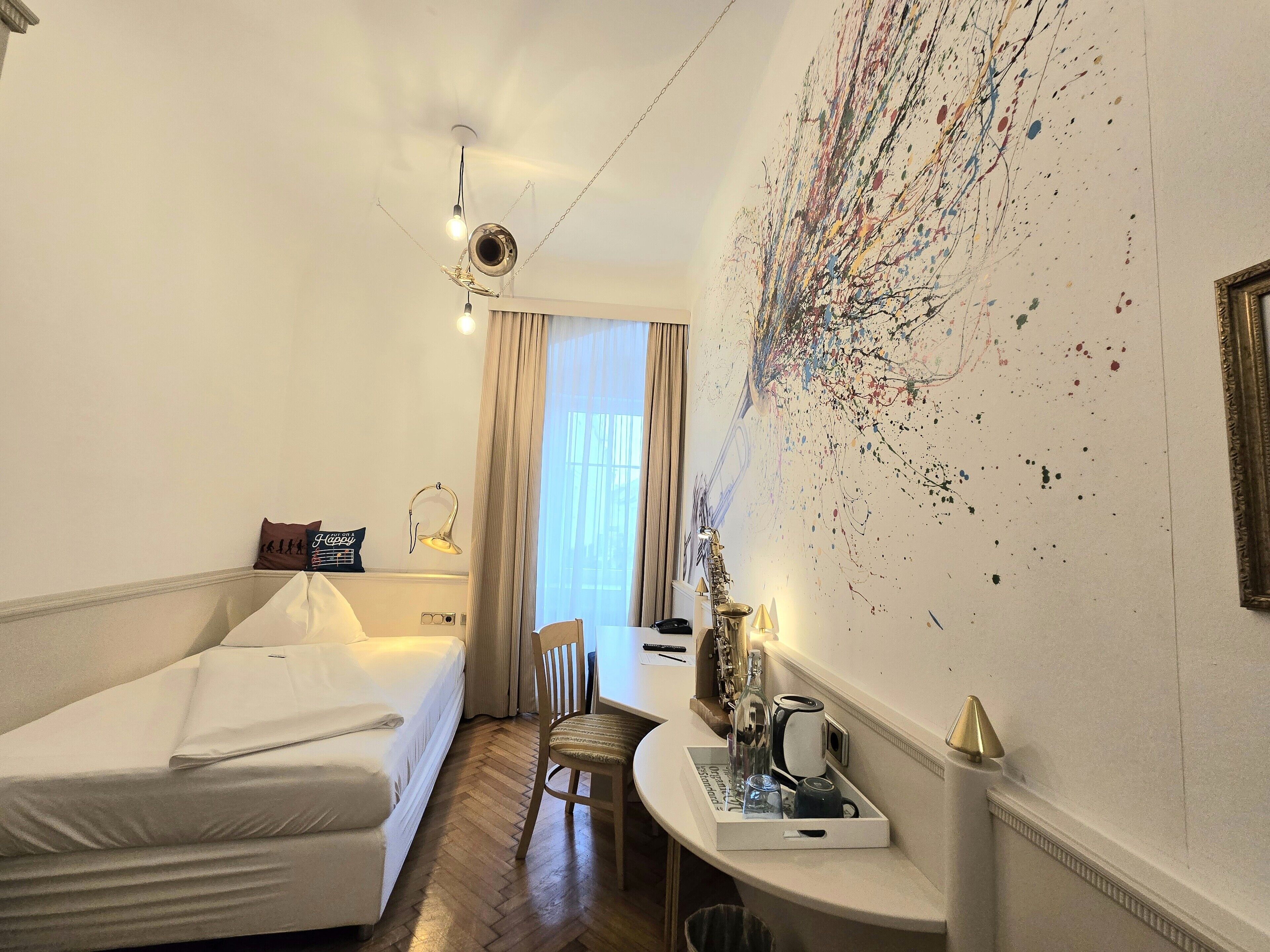 Habitación de hotel con cama, una silla, una mesita con una bandeja de objetos, una pared con un cuadro abstracto colorido y una ventana con cortinas.