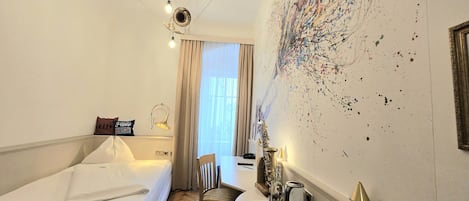 Habitación de hotel con cama, una silla, una mesita con una bandeja de objetos, una pared con un cuadro abstracto colorido y una ventana con cortinas.
