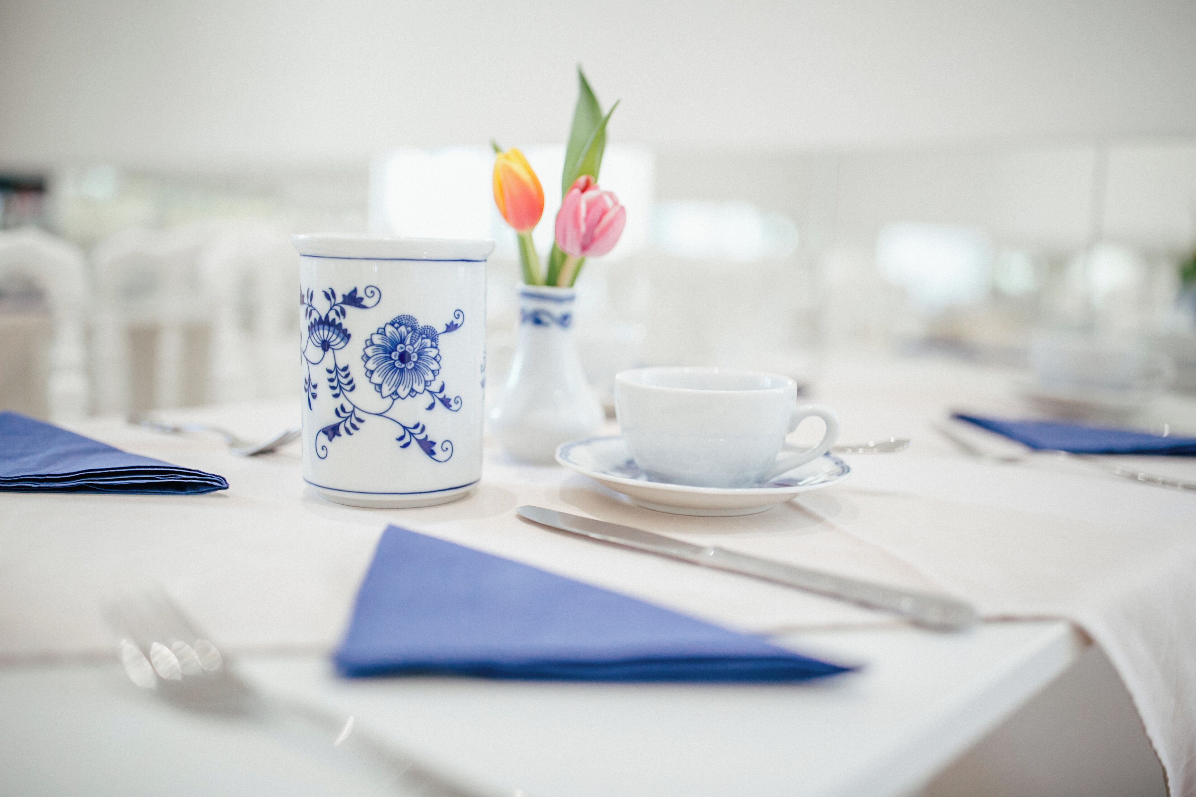 A table set with a white tablecloth, a blue and white floral mug, a white cup and saucer, a vase with tulips, and a blue napkin.
