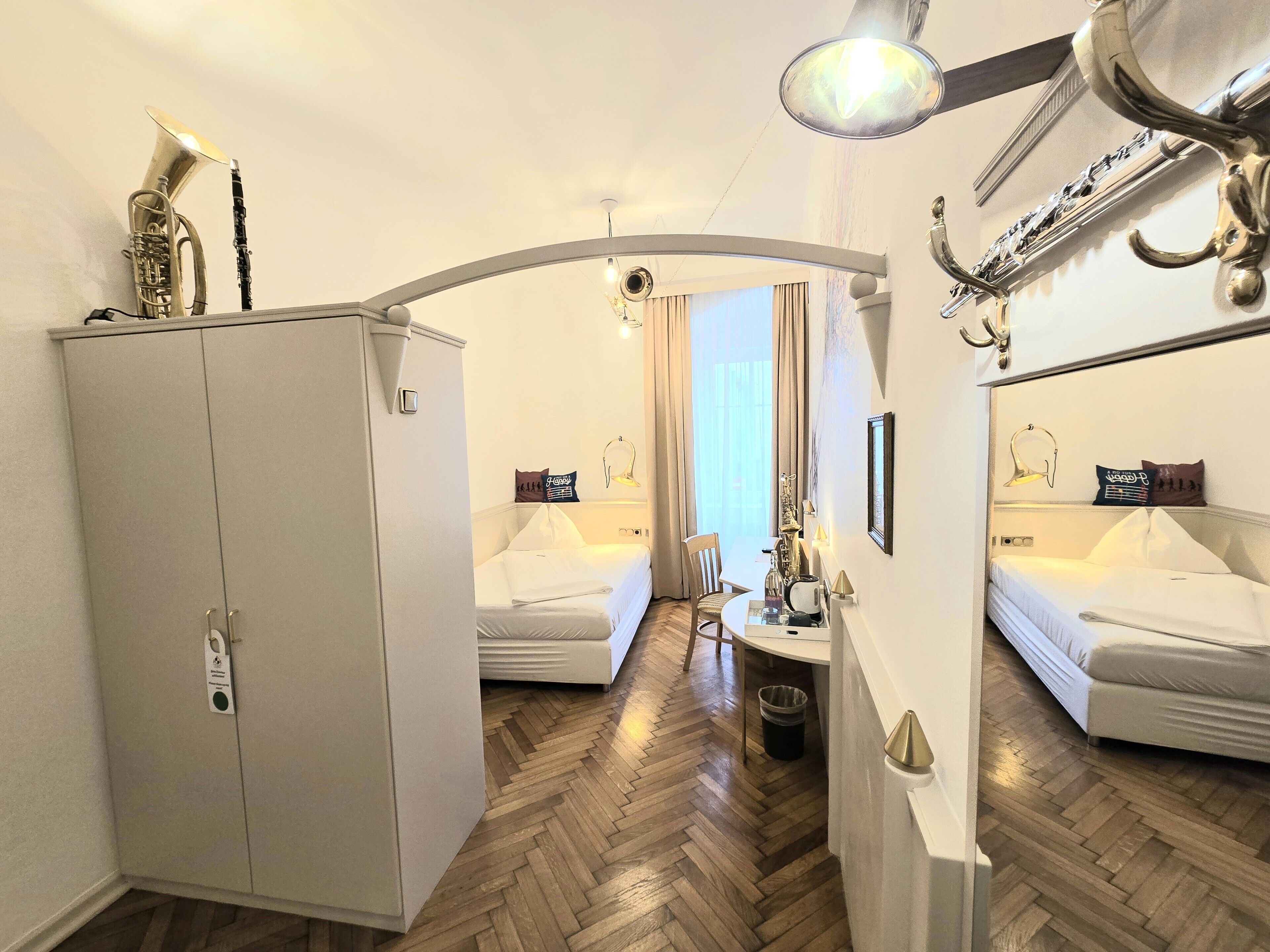 A hotel room with a mirrored wardrobe, a bed, a small table with a lamp, and a window with curtains.