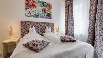 A bedroom with a large bed, two bedside tables with lamps, a floral artwork above the bed, and patterned curtains.