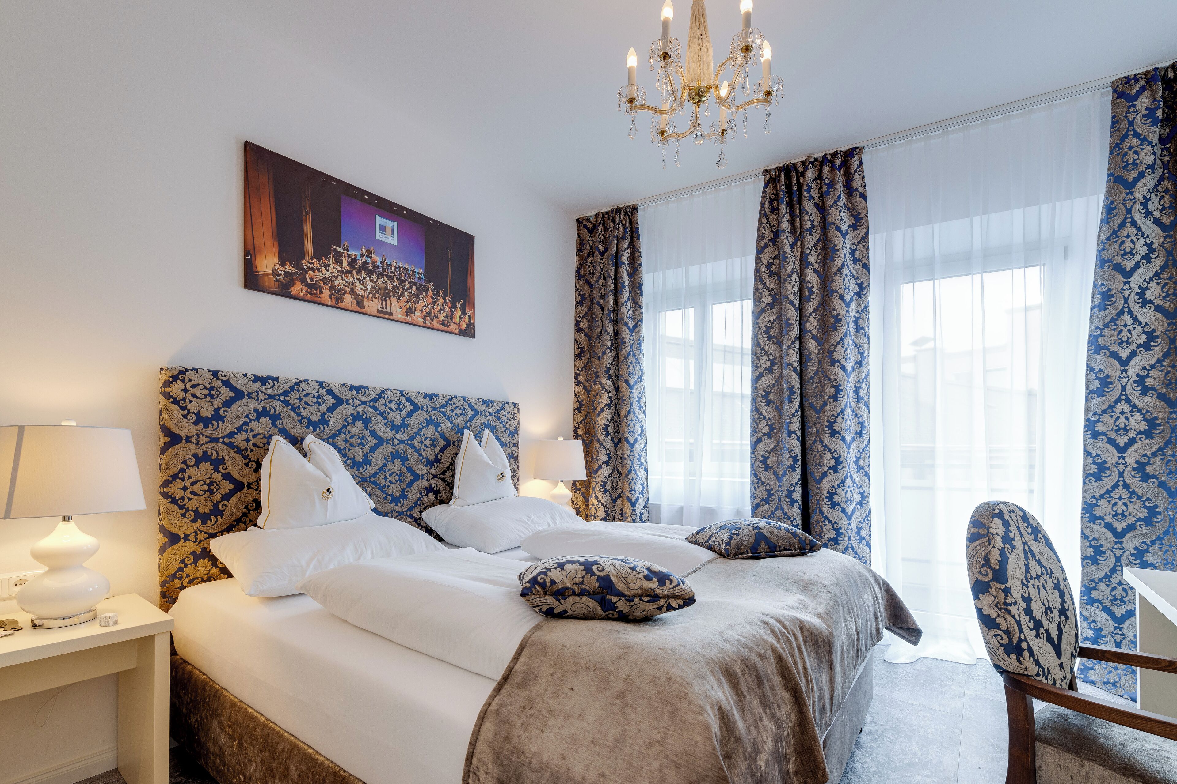 A hotel room with a large bed, a desk, a chair, a chandelier, and a painting on the wall.