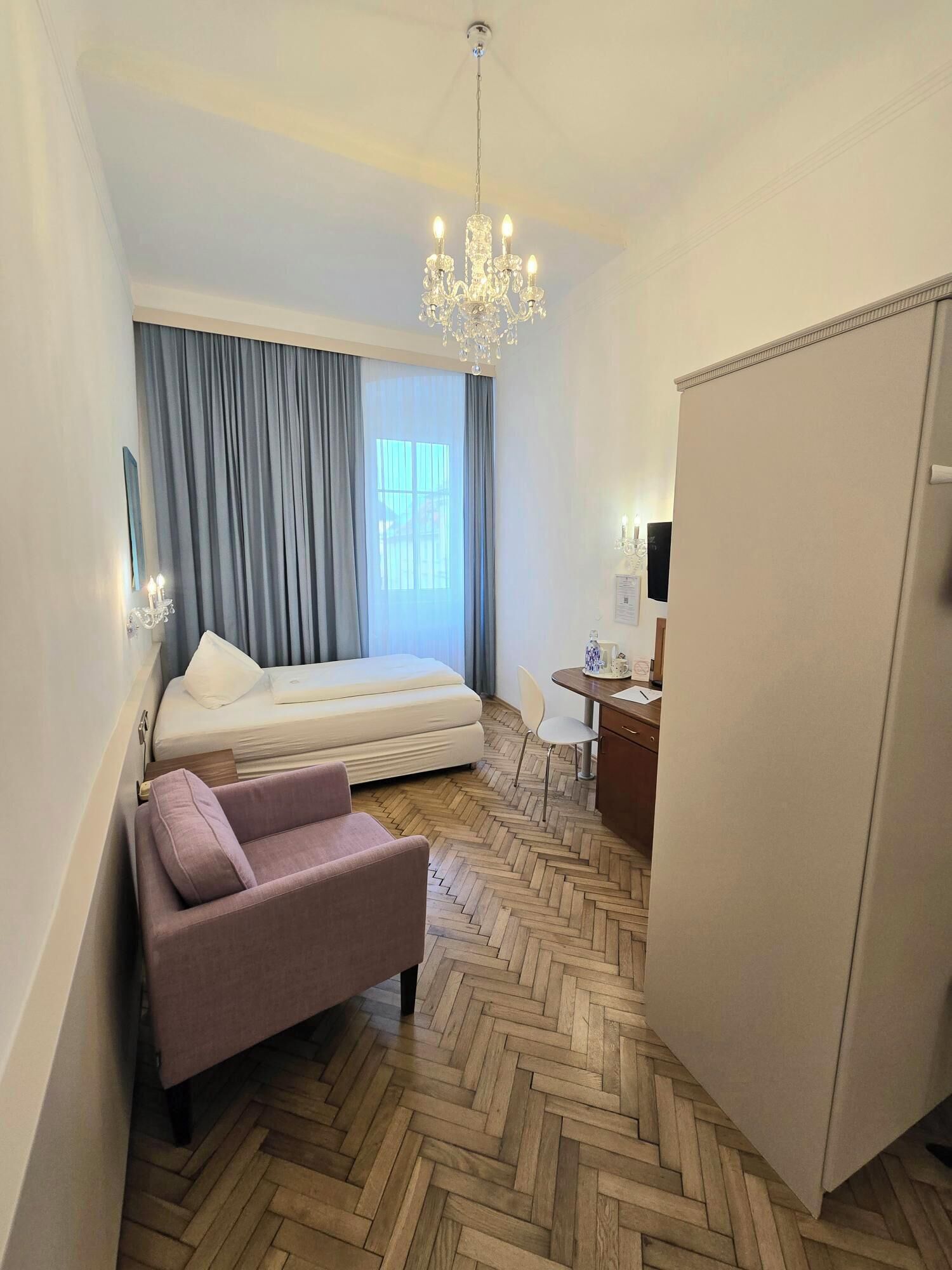 A hotel room with a bed, a desk, a chair, a wardrobe, a chandelier, and a window with curtains.