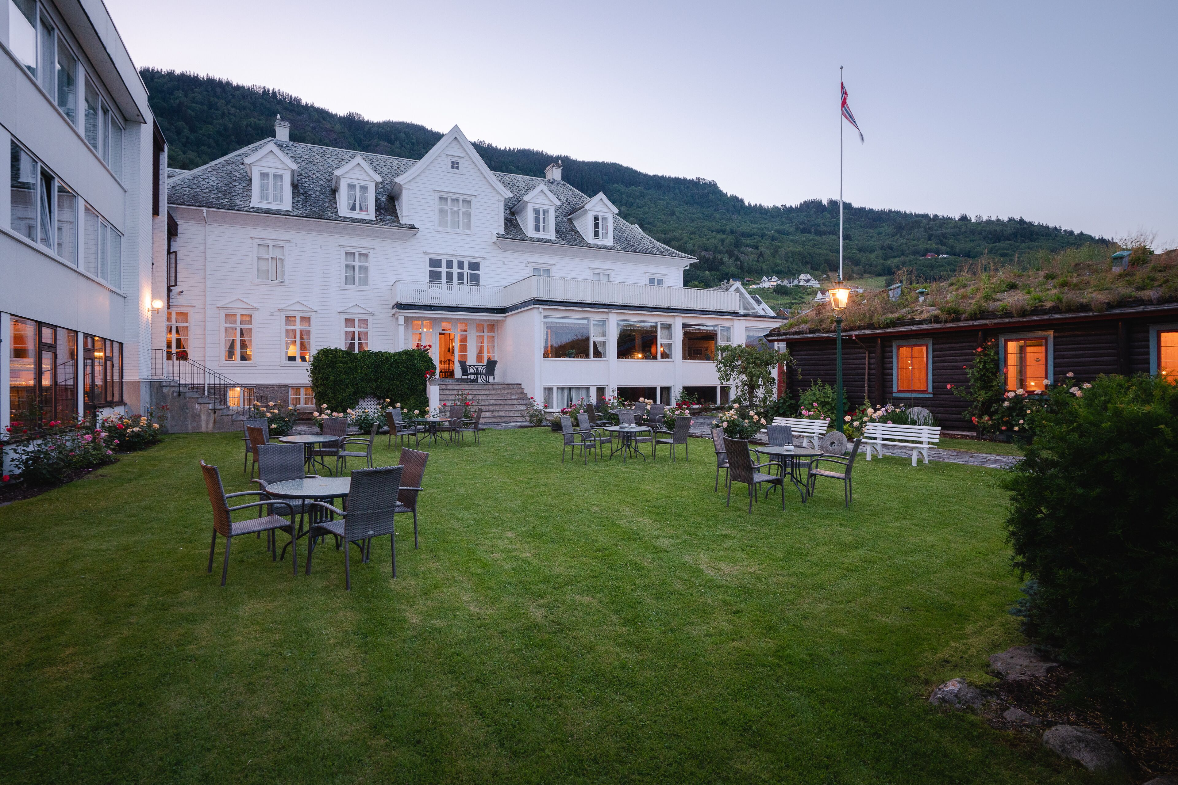 A large white building with multiple windows and a flag, surrounded by a well-maintained lawn and outdoor seating area with tables and chairs.