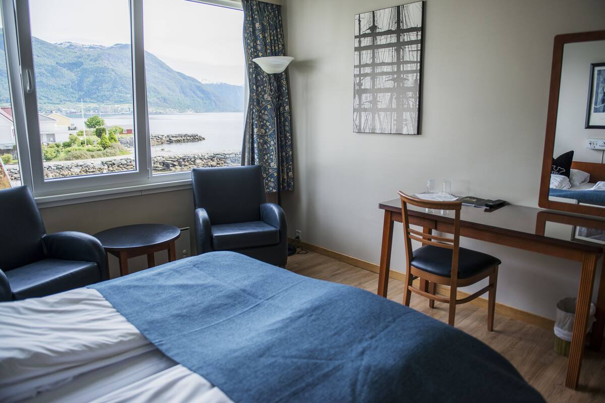 A hotel room with a bed, two chairs, a desk, and a view of a lake and mountains.