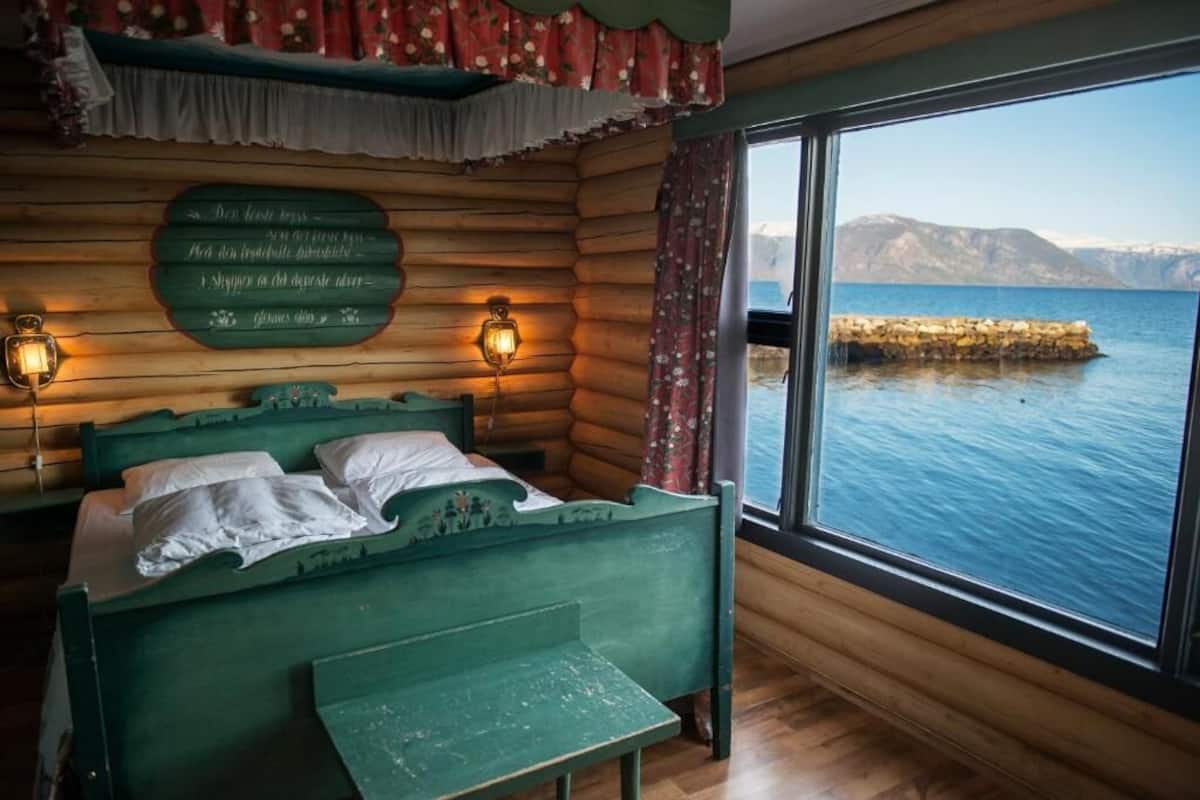 A cabin-style room with a bed, a bench, and a large window overlooking a body of water and mountains.
