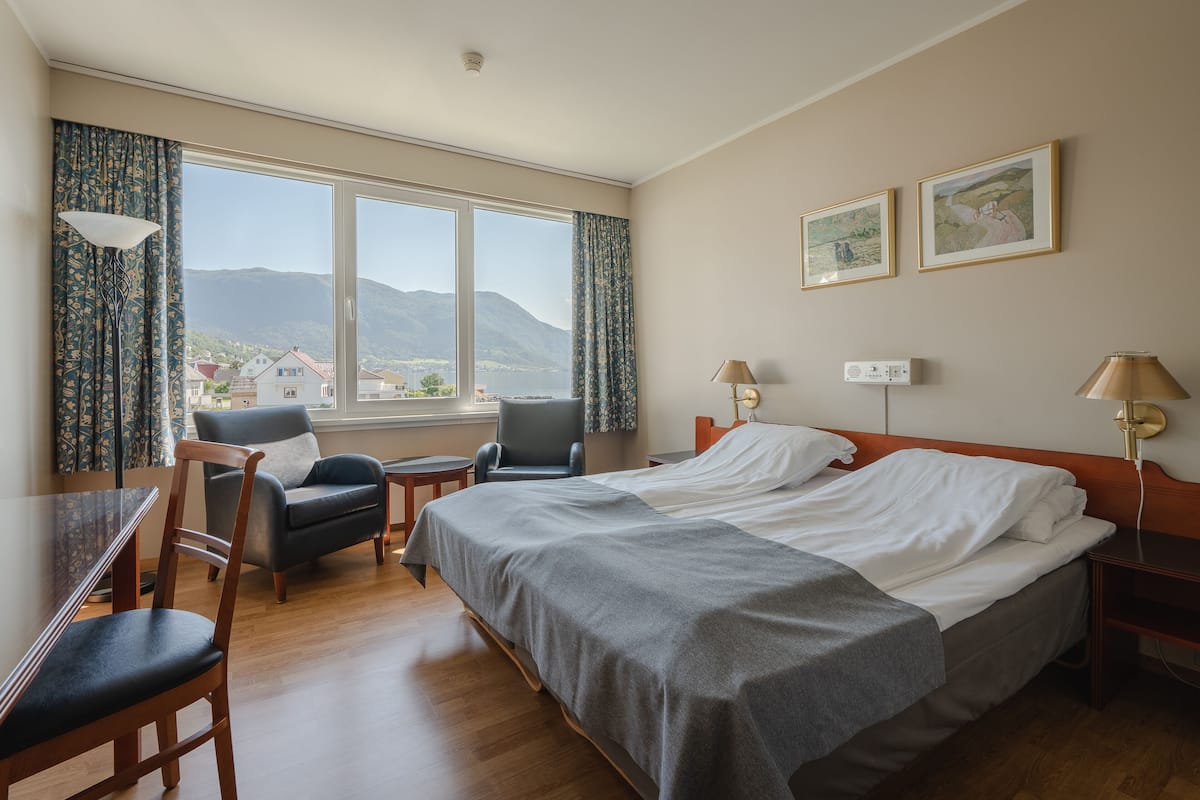 A hotel room with a bed, a desk, a chair, a window with curtains, and two wall-mounted lamps.