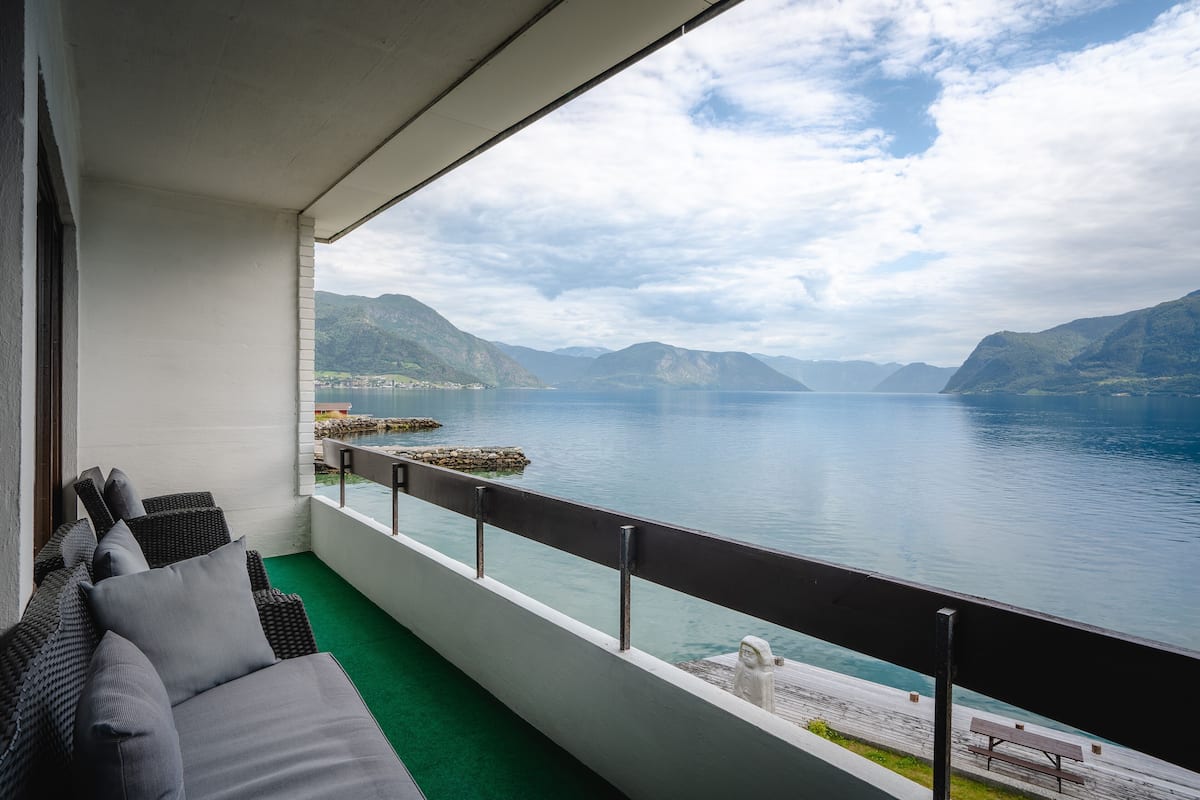 A balcony with a view of a calm lake, mountains, and a wooden dock.