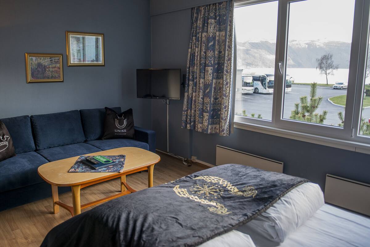 A hotel room with a dark blue sofa, a wooden coffee table, a bed with a dark blanket, a flat-screen TV, and a window with a view of a parking lot and mountains.
