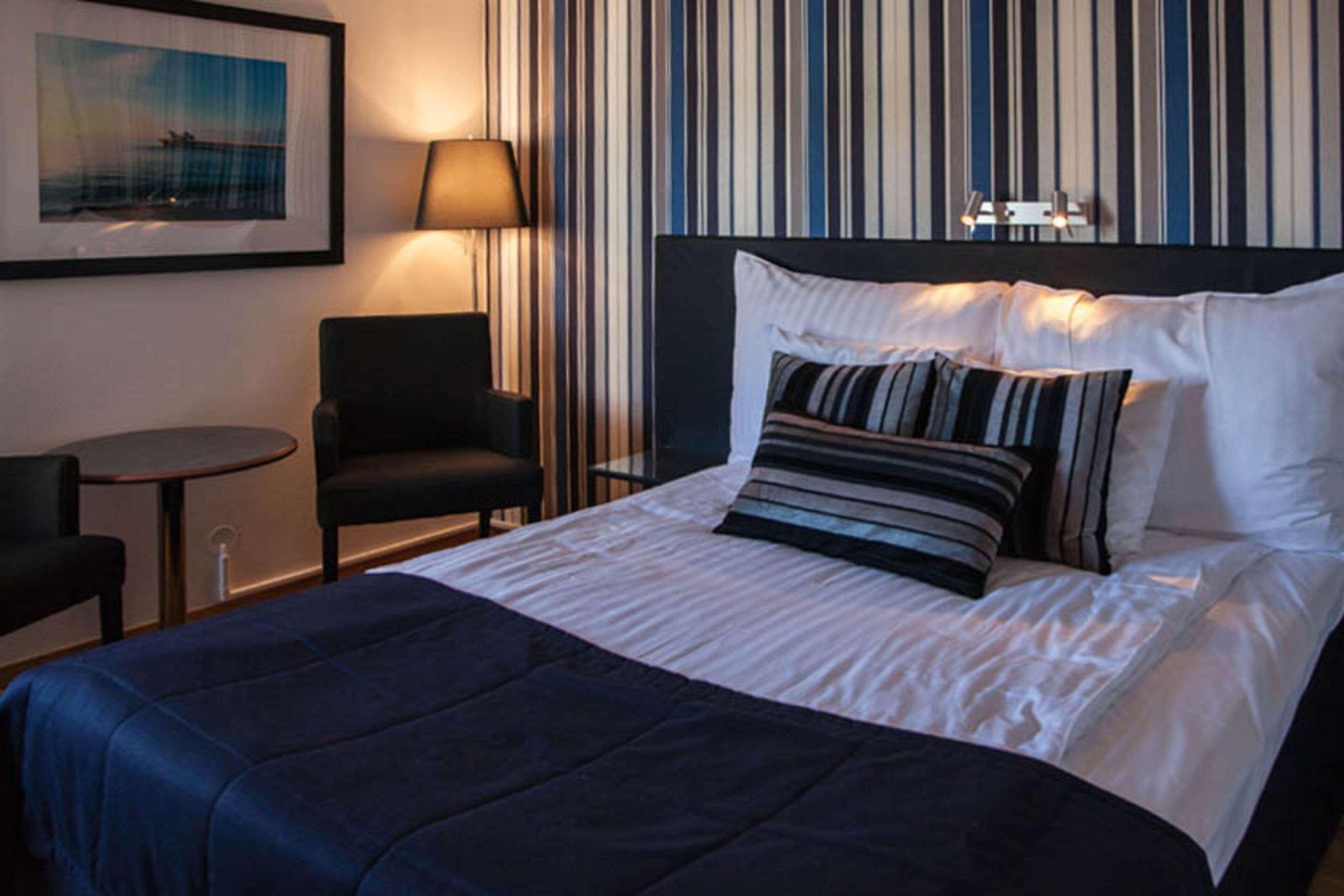 Habitación de hotel con cama, una silla, una mesita pequeña, una lámpara y un muro a rayas.