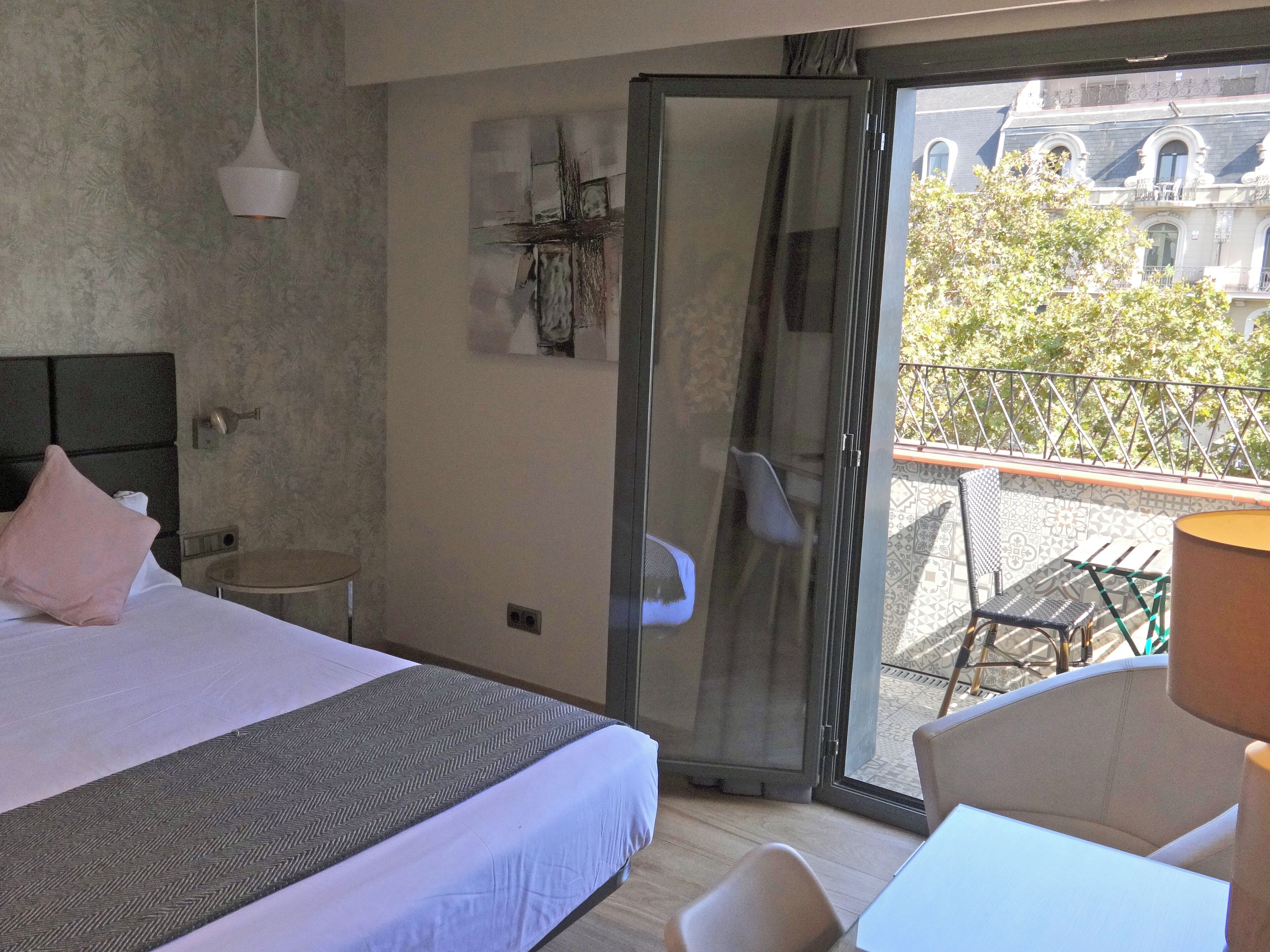Une chambre d’hôtel avec un lit, une petite table, une chaise et un balcon donnant sur des immeubles et des arbres.