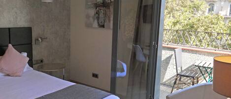 Une chambre d’hôtel avec un lit, une petite table, une chaise et un balcon donnant sur des immeubles et des arbres.
