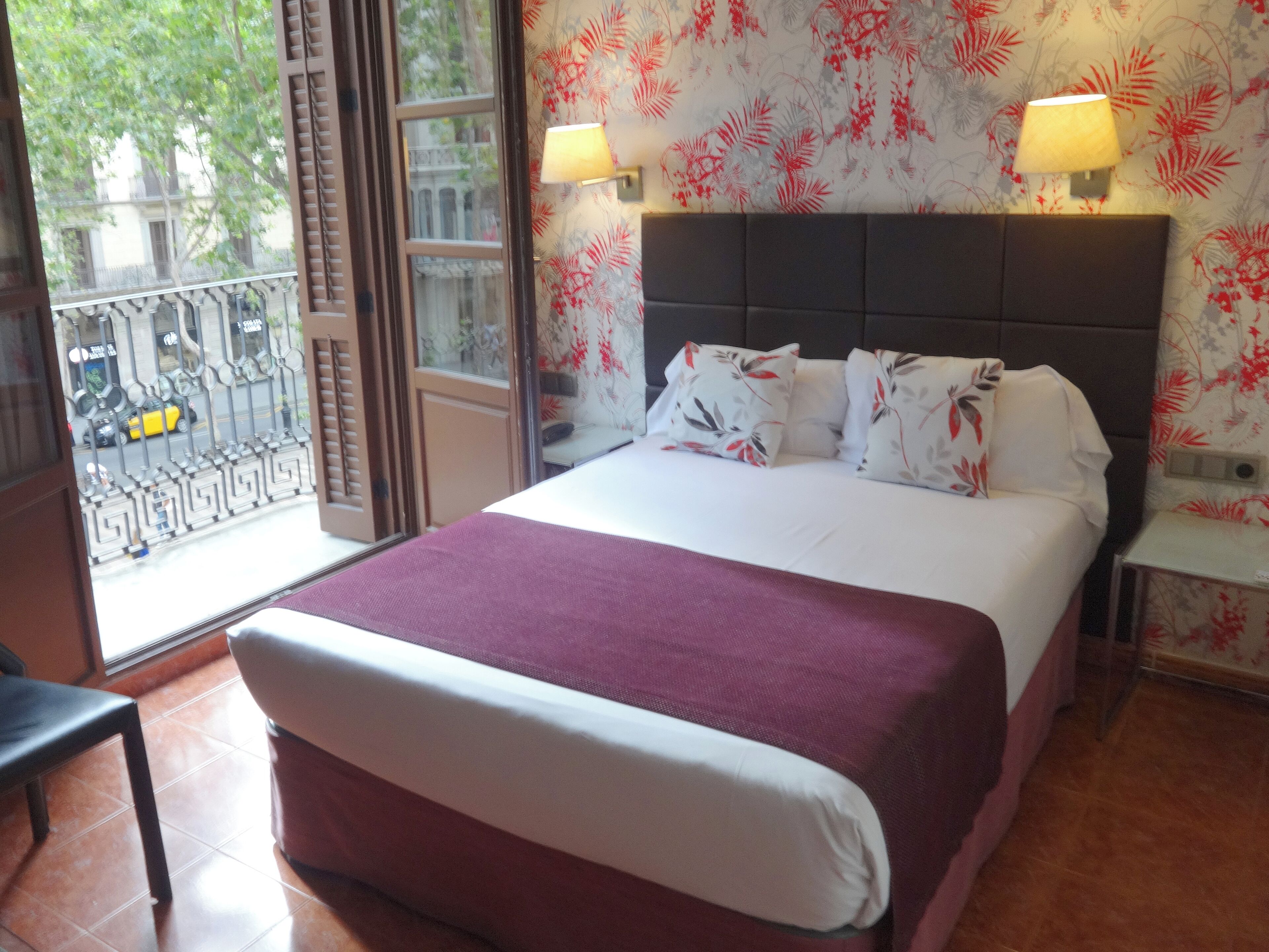 Une chambre d’hôtel avec un lit, deux lampes de chevet, une fenêtre donnant sur la rue et un papier peint à motifs.