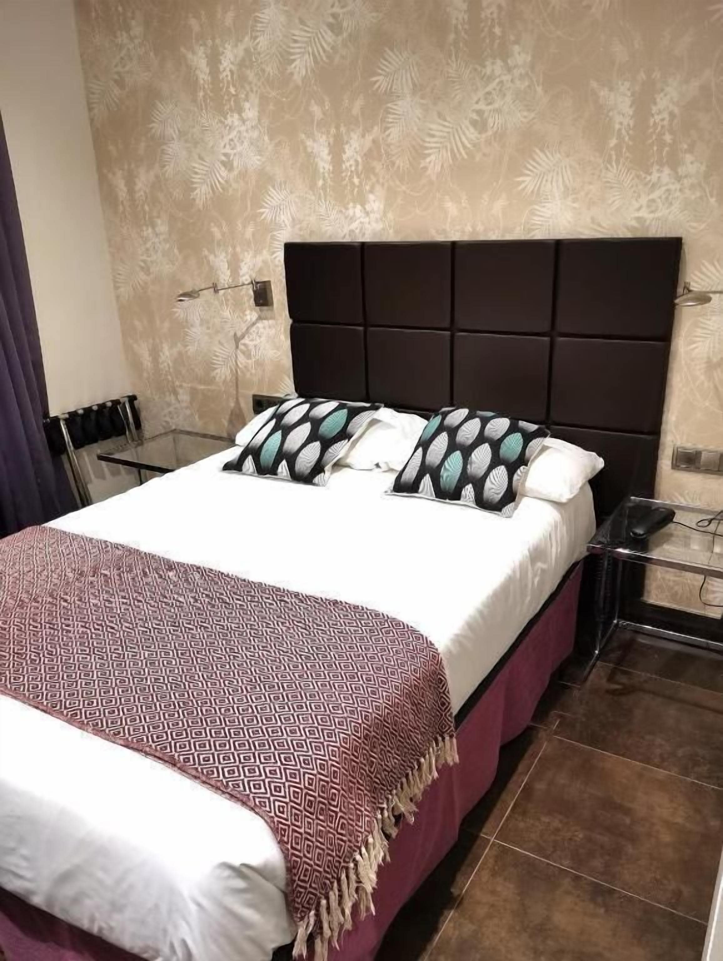 A bed with a patterned bedspread, a dark headboard, and a patterned wall behind it.