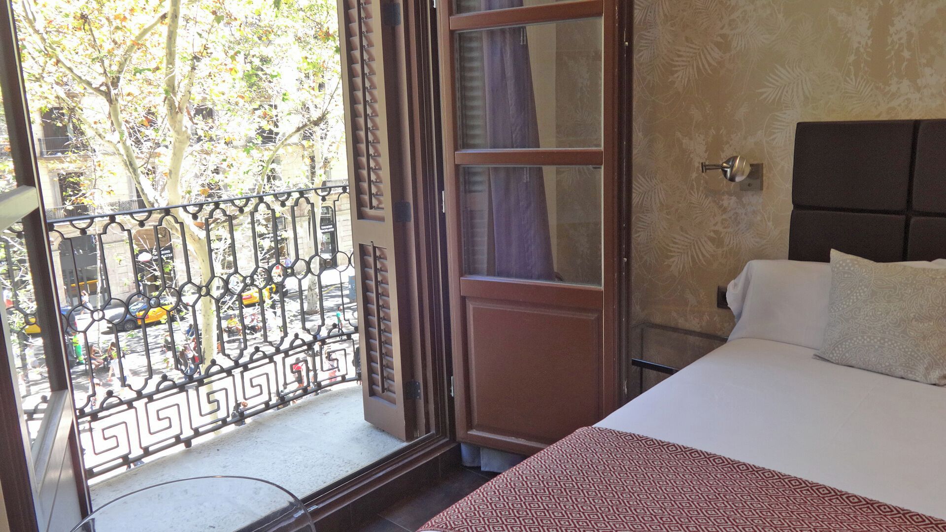 A hotel room with a bed, a window with a view of the street, and a balcony with a decorative railing.