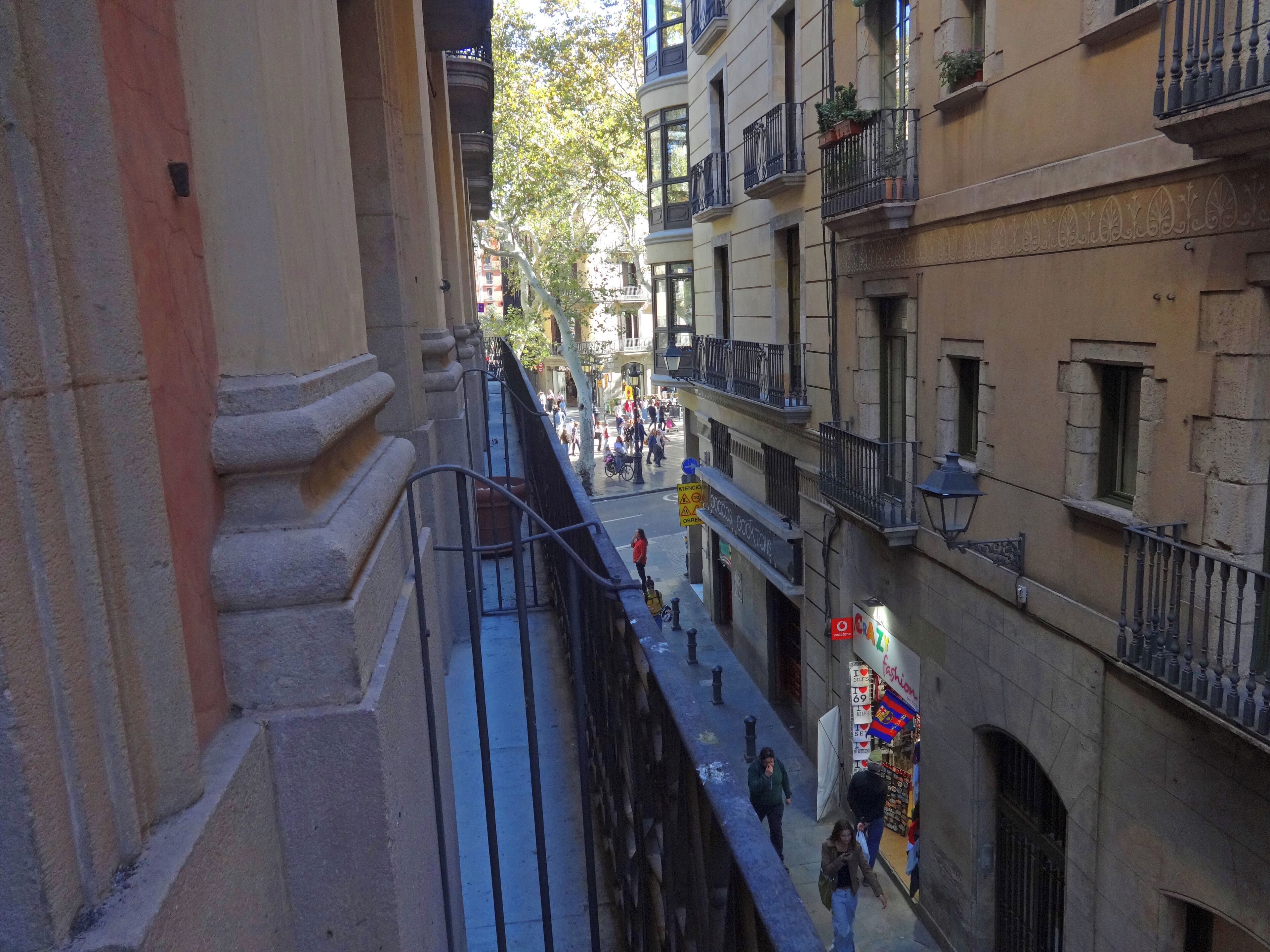 A narrow urban street with buildings on either side, balconies, and pedestrians.