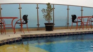 A pool area with a glass balustrade, three red chairs, and a potted plant. - Le Royal Corniche (Salmiya)