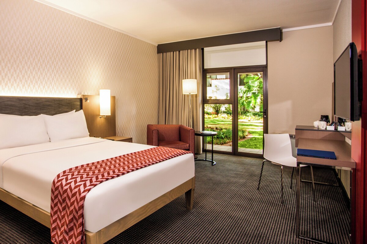 Habitación de hotel con cama, escritorio, silla, lámpara y vistas al exterior.