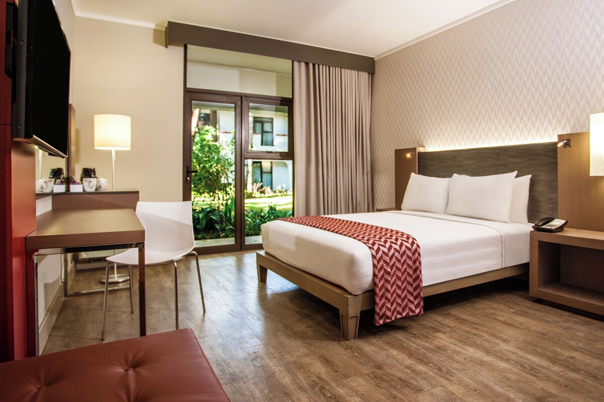 Een hotelkamer met een bed, nachtkastje, lamp, stoel en uitzicht op het omliggende landschap.