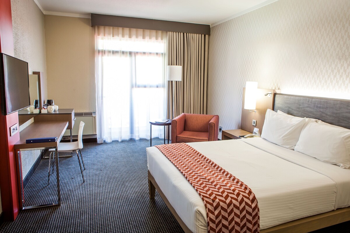 Quarto de hotel com cama, escrivaninha, cadeira, televisão e janela com cortinas.