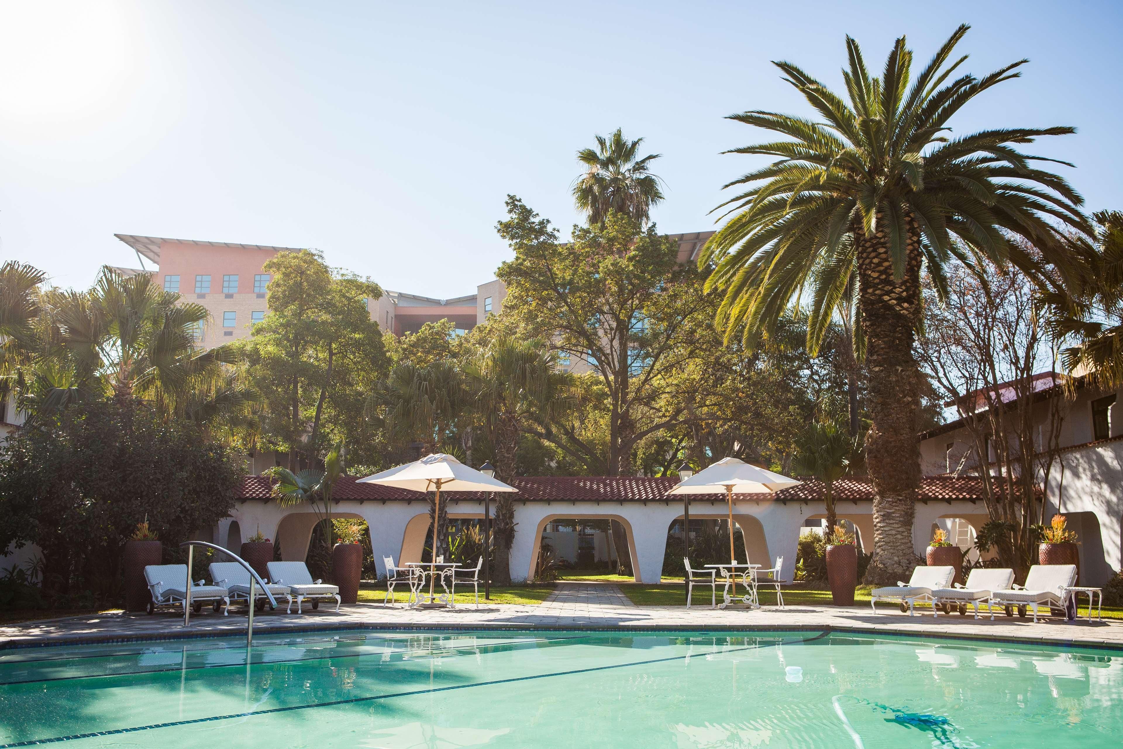 Un área de piscina con sillas de descanso y sombrillas, rodeada de palmeras y un edificio con techo de tejas.