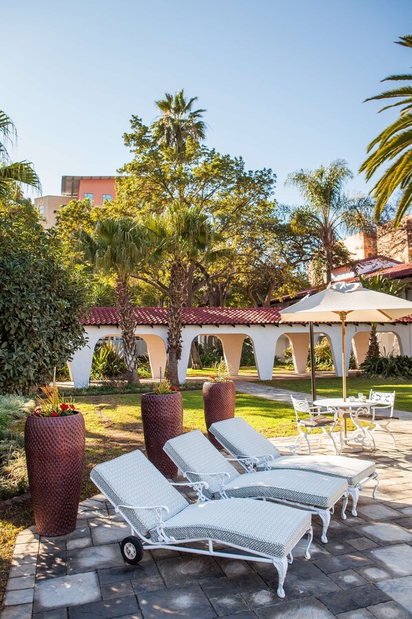 Een terras met lounge stoelen, een pergola en een pad dat naar een gebouw met een rode pannendak leidt.