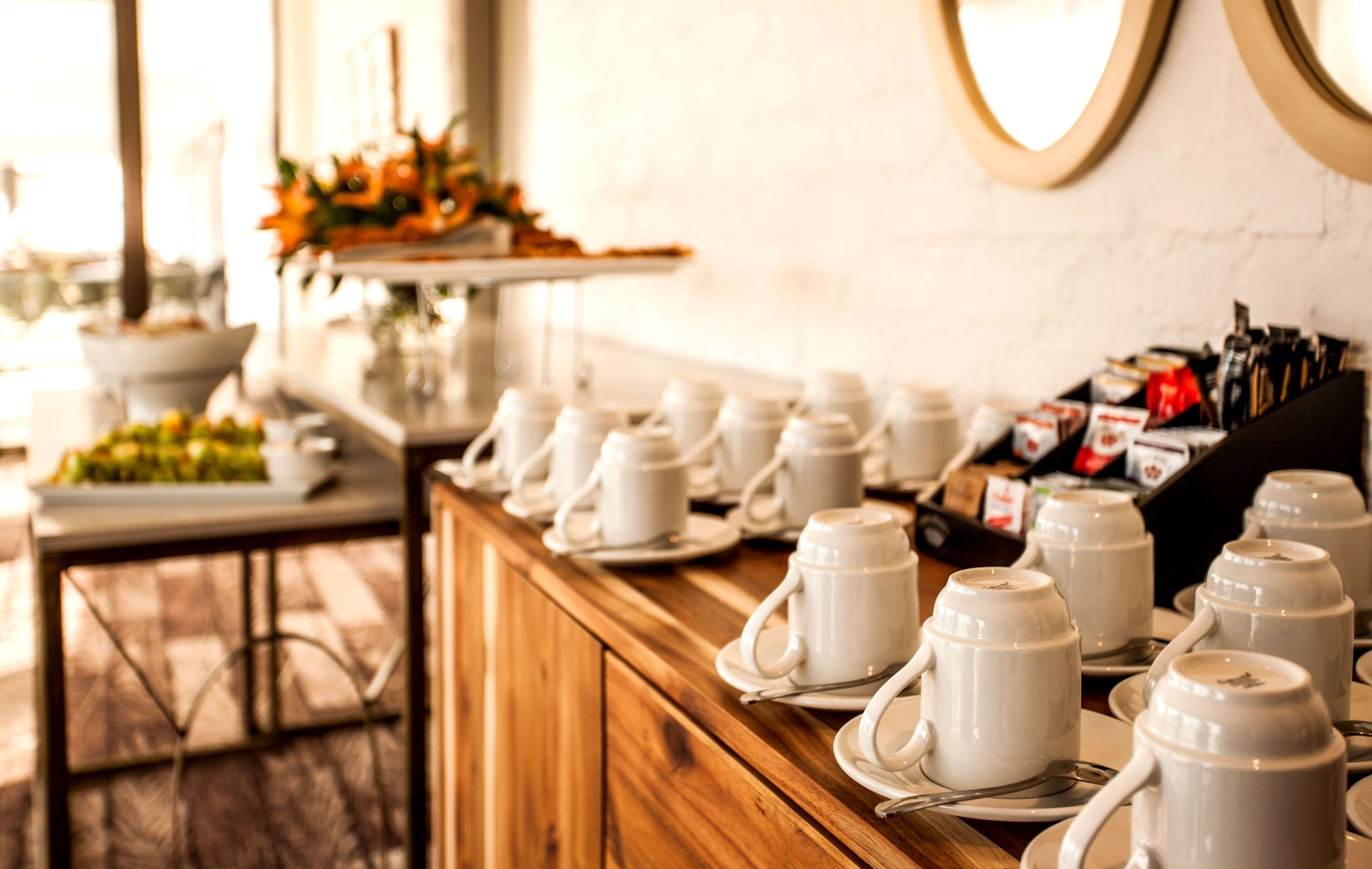 A buffet setup with white ceramic mugs and plates on a wooden counter, a floral arrangement, and a white brick wall.