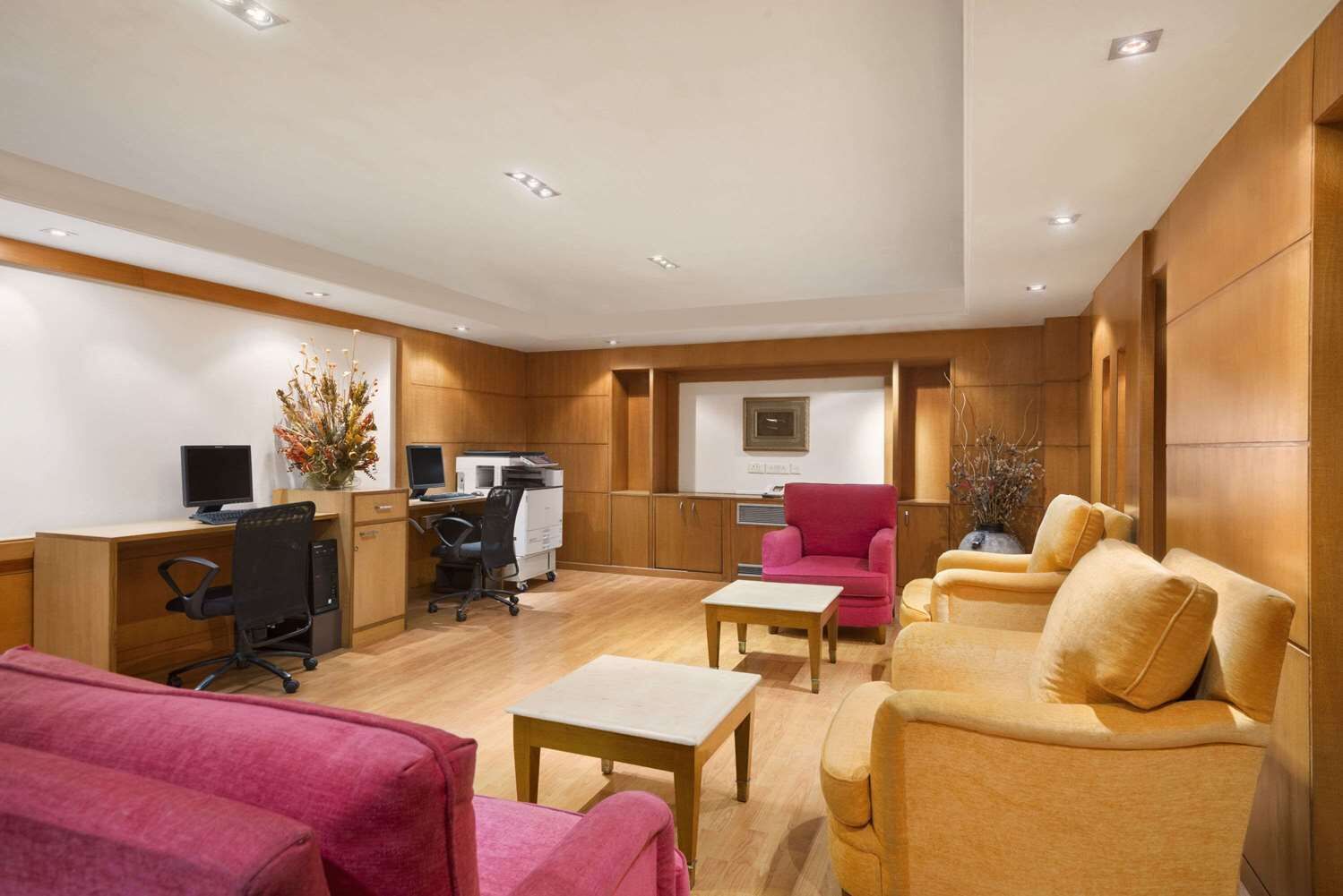 A modern hotel room with a wooden floor, a desk with a computer, a red chair, a yellow sofa, and a small table.