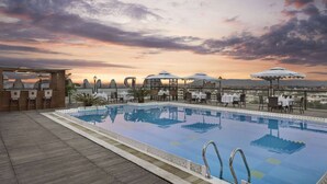 Una piscina en la azotea con vistas a la ciudad al atardecer, rodeada de asientos exteriores y sombrillas.