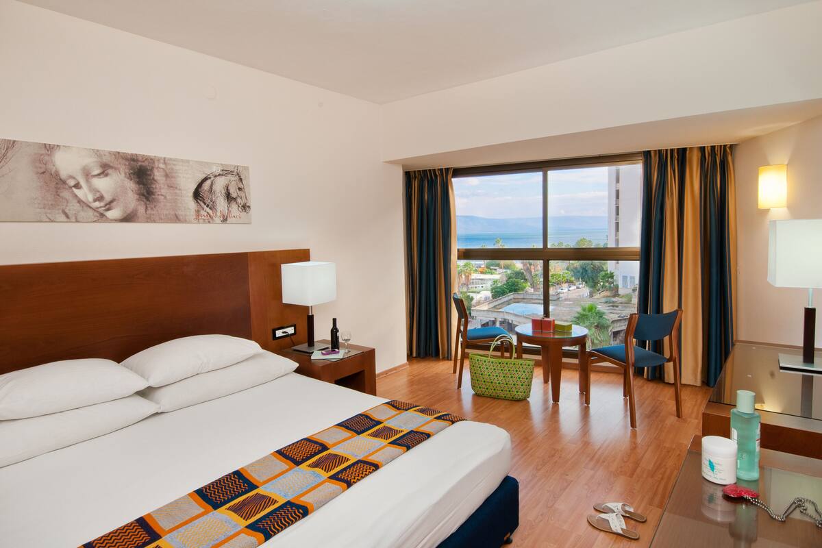 Une chambre d’hôtel avec un lit, un bureau, une chaise, une fenêtre donnant sur un beau paysage et un tableau accroché au mur.