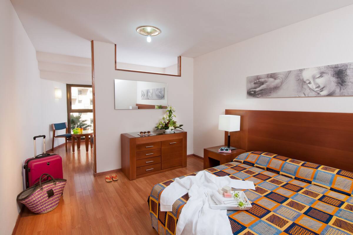 Une chambre d’hôtel avec un lit, une commode, un bureau, une chaise, une valise et un tableau au mur.