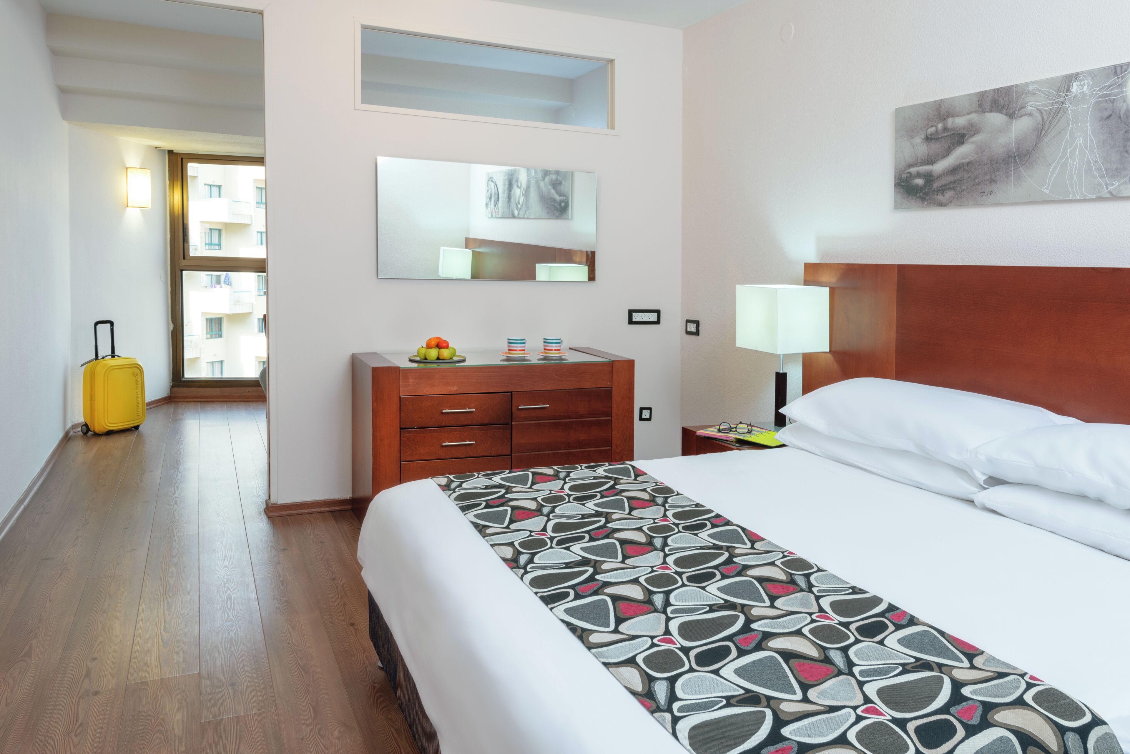 Una habitación de hotel con cama, cómoda, maleta, espejo y un cuadro en la pared.