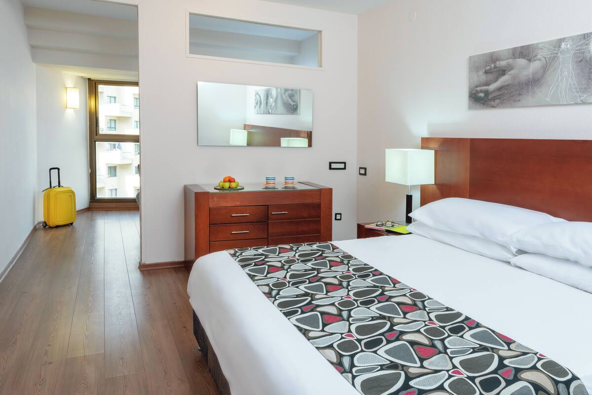 Une chambre d’hôtel avec un lit, une commode, une valise, un miroir et une fenêtre.