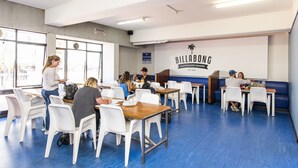 En spiseplads med hvide stole og borde, en væg med skilt der står "BILLABONG BACKPACKERS RESORT EST 2000", og store vinduer der lukker naturligt lys ind.