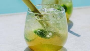 Two glasses of a yellow beverage with mint leaves and a straw, placed on a white surface with a pool in the background.