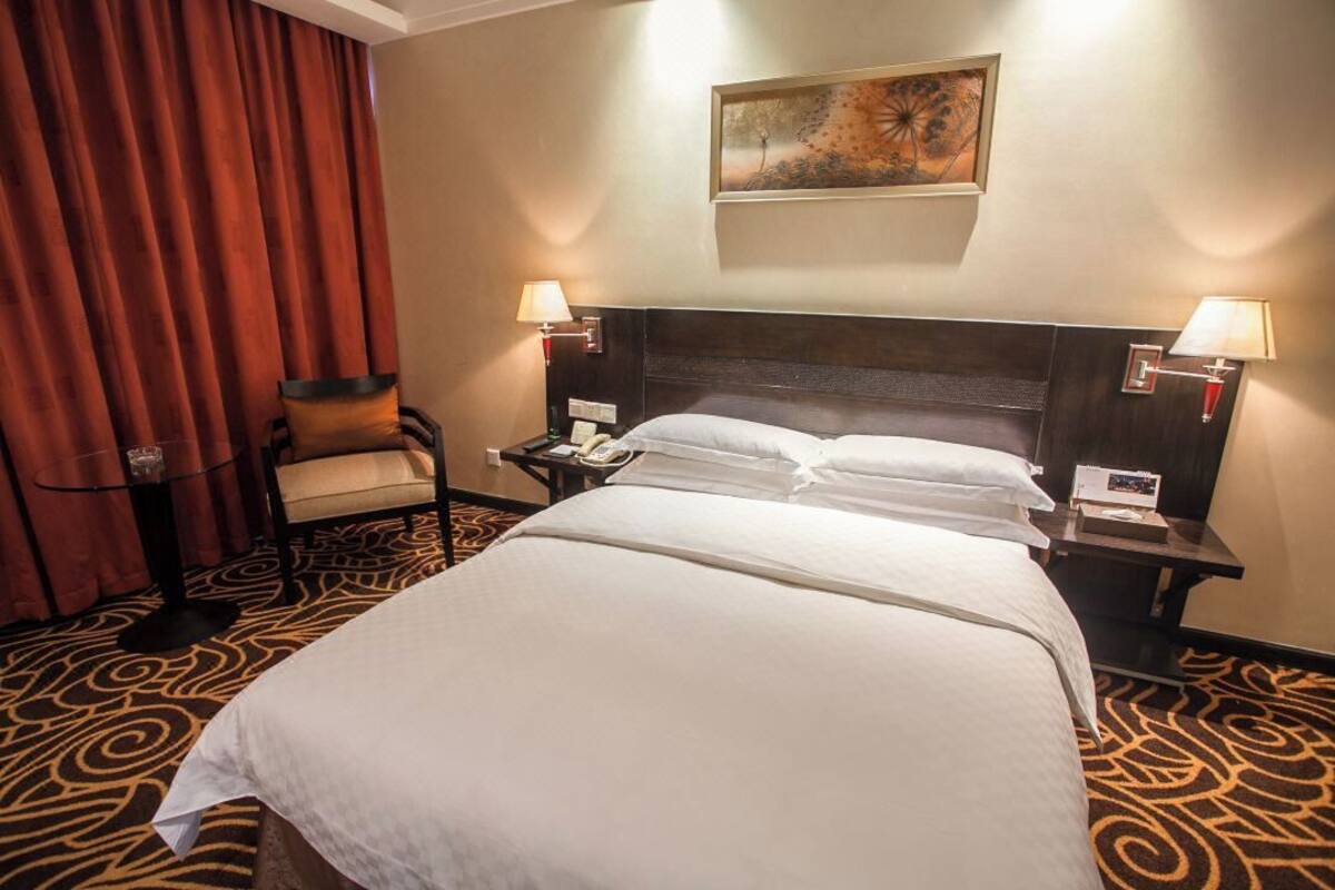 A hotel room with a large bed, two bedside tables with lamps, a chair, a red curtain, and a painting on the wall.