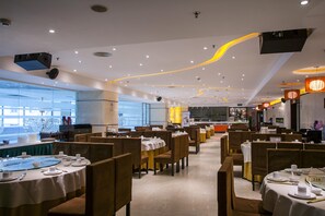 A restaurant with multiple dining tables and booths, a kitchen in the background, and a view of the outside through large windows. - Huashi Hotel (Guangzhou)