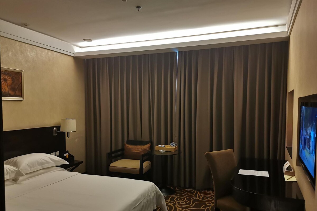 Ein Hotelzimmer mit einem Bett, einem Schreibtisch, einem Sessel, einem Fernseher und einem Vorhang.