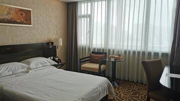 Una habitación de hotel con una cama grande, una silla, una mesita pequeña y una ventana con cortinas traslúcidas.