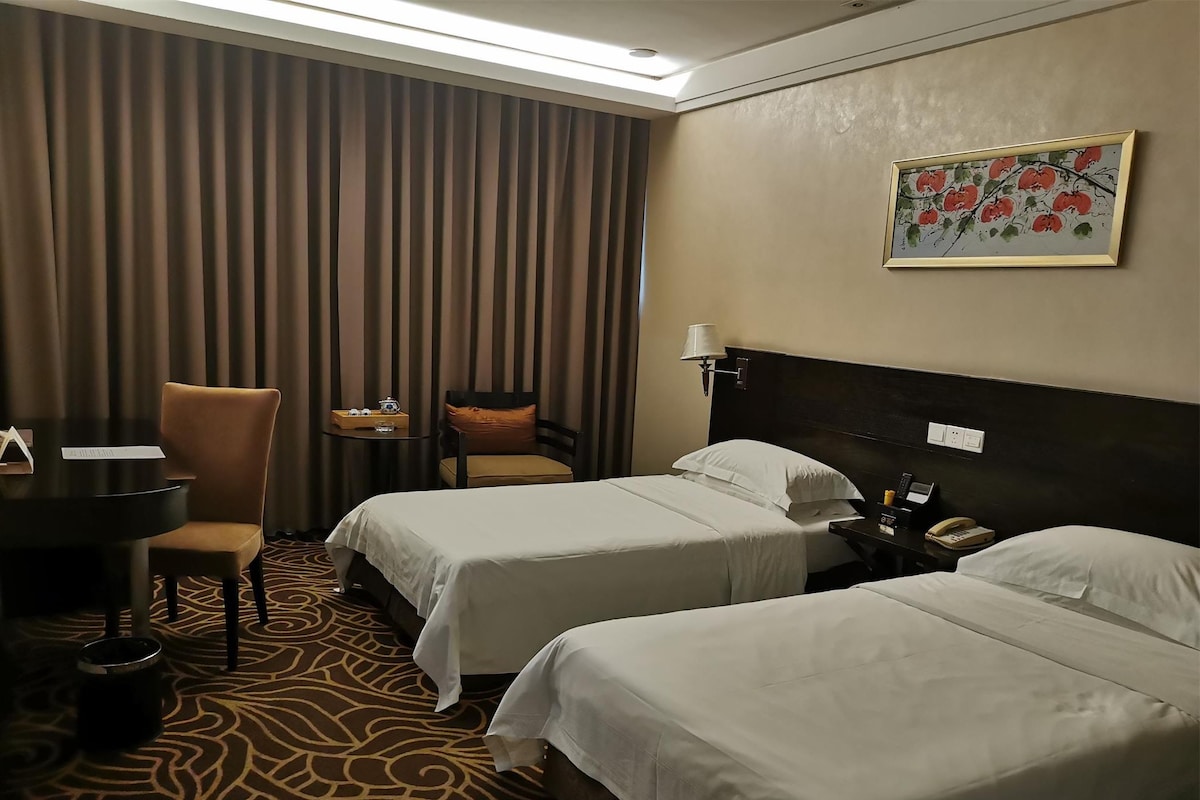 Ein Hotelzimmer mit zwei Betten, einem Schreibtisch, einem Stuhl, einem kleinen Tisch und einem Bild an der Wand.