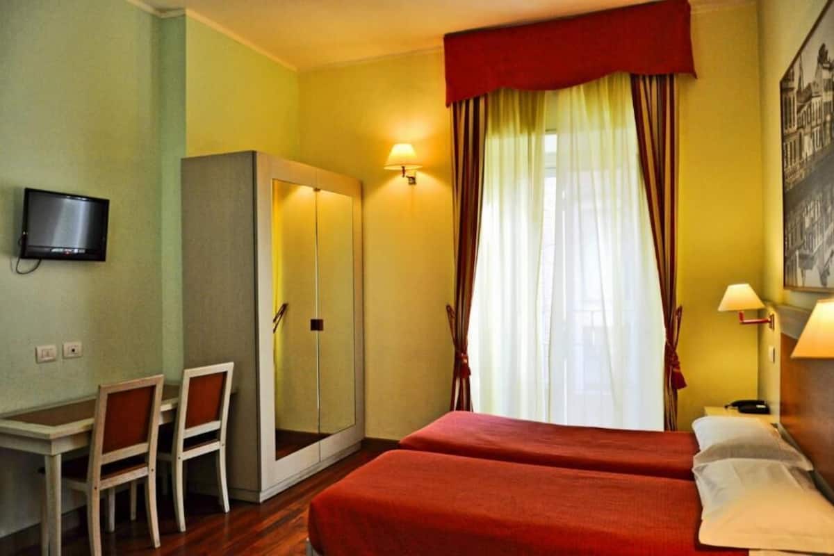 A hotel room with a bed, a desk with chairs, a TV, a wardrobe, and a window with curtains.