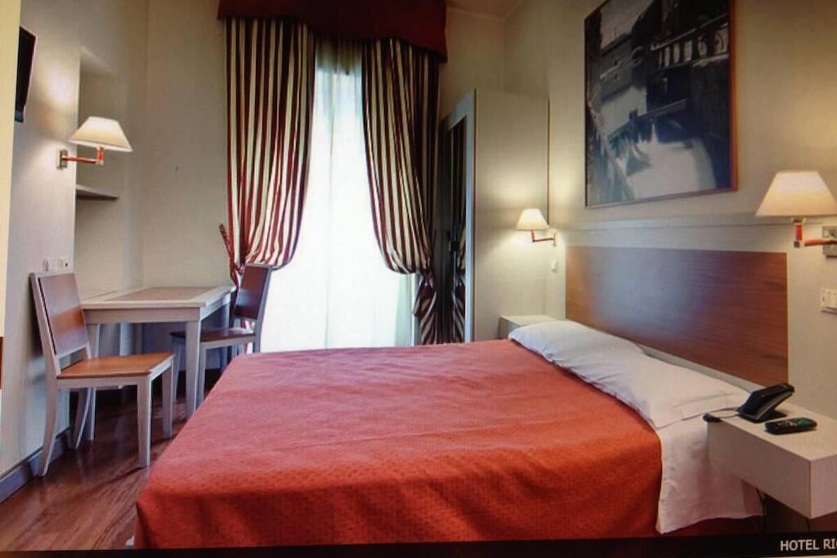 A hotel room with a large bed, a desk, two chairs, a lamp, and a painting on the wall.