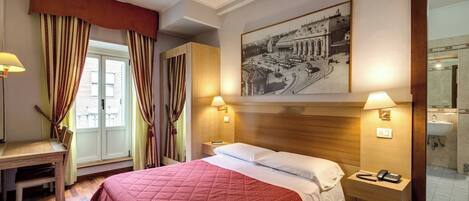 Une chambre d’hôtel comprenant un lit, des tables de chevet, un bureau, une chaise, une fenêtre avec des rideaux, une lampe fixée au mur et un tableau encadré.
