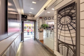 Un pasillo con una pared de vidrio decorada con diseños de metalería, una zona de recepción con un reloj y una señal de salida visible.