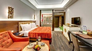 Hotelkamer met een bed, een slaapbank, een bureau, een televisie en een eettafel met fruit en een fles.